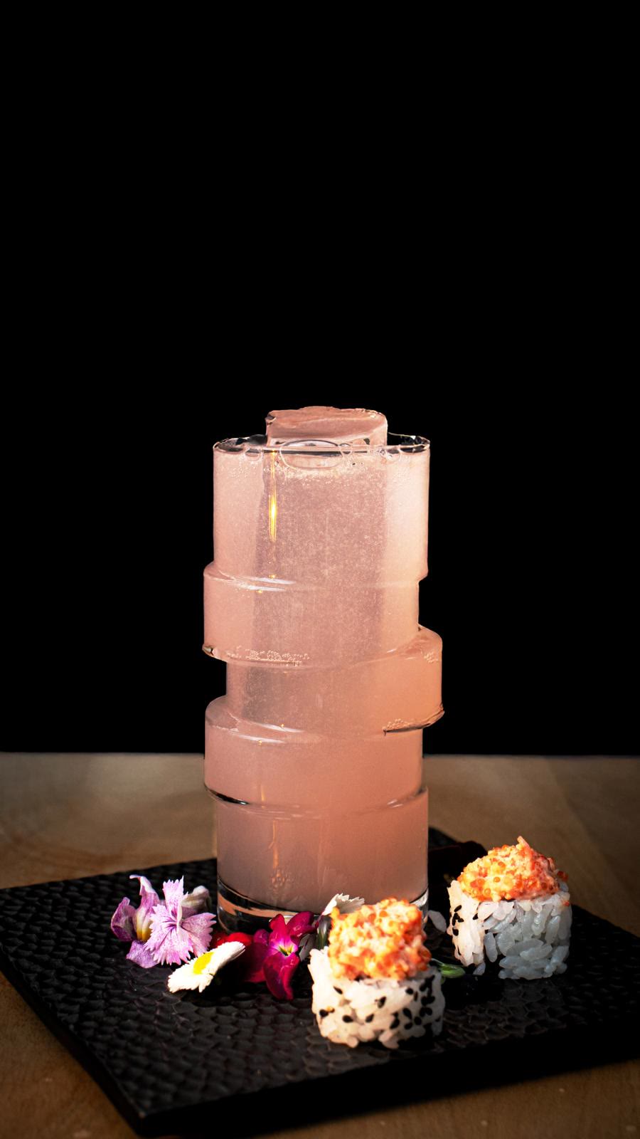 Cóctel rosado con hielo tallado junto a piezas de sushi
